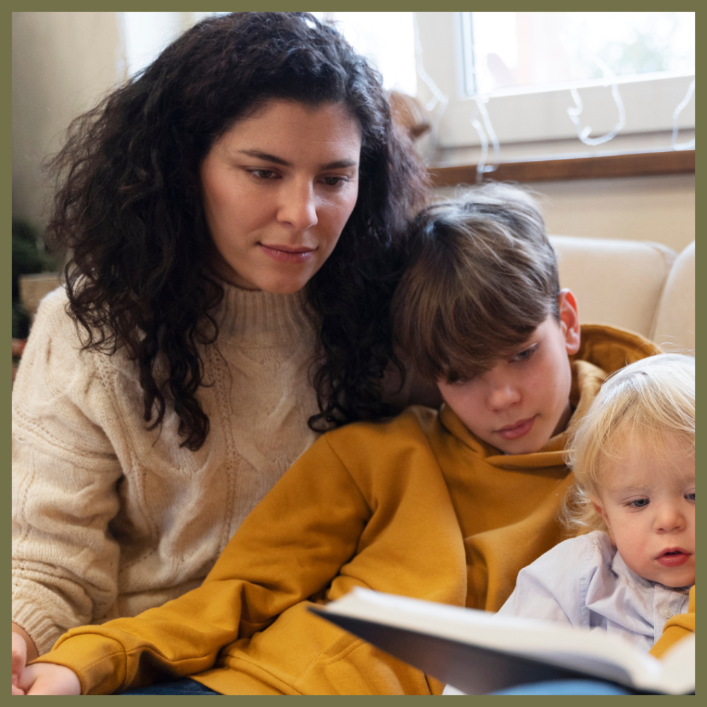 A woman with curly dark hair sits closely with two children, a boy in a yellow hoodie and a younger blonde child, all looking down intently, at a book.